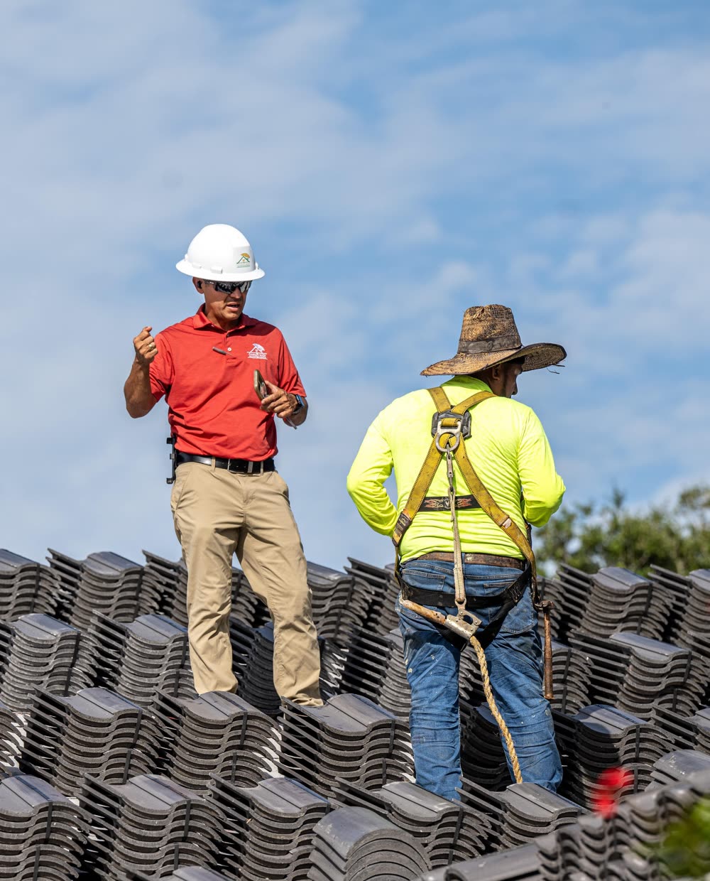 Campany foreman directing a harnessed crew member atop stacked roof tile, fall-protection gear in use